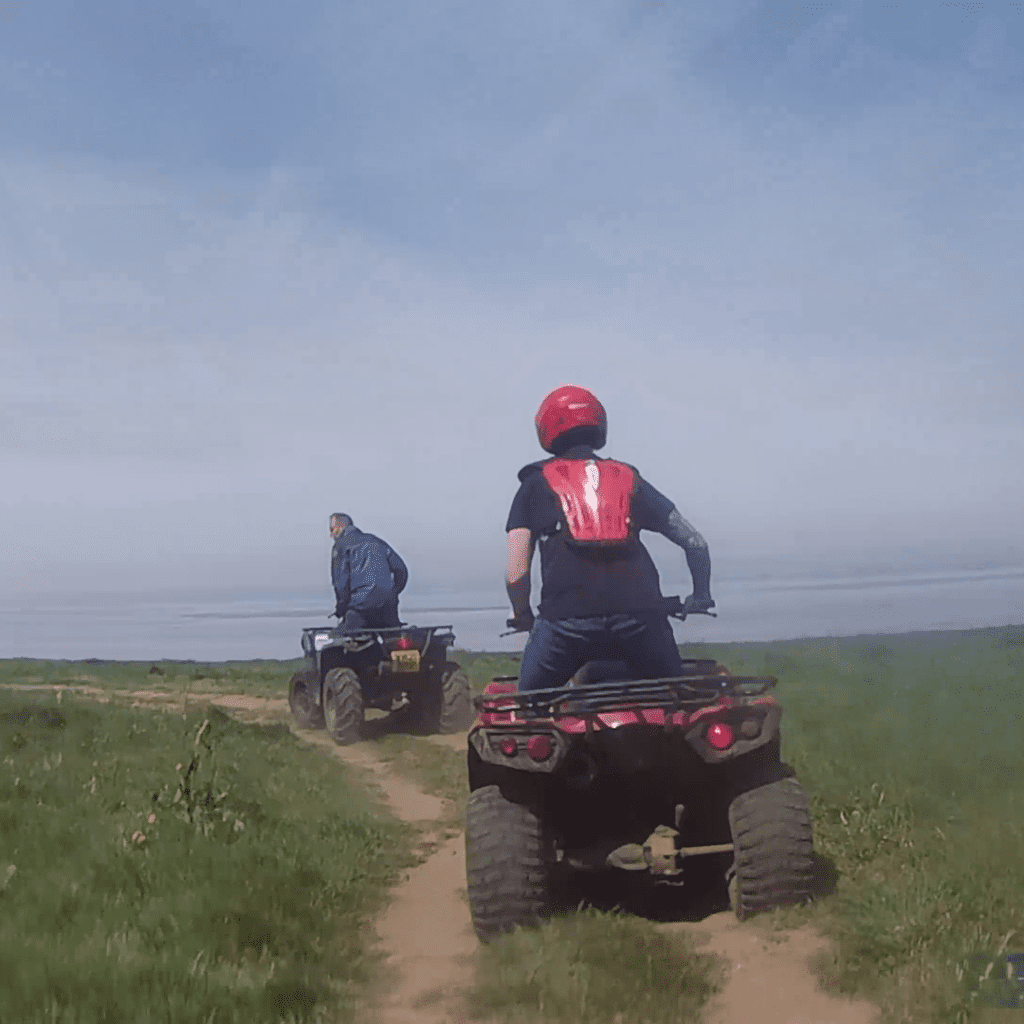 Two people riding all-terrain vehicles (ATVs) on a dirt path through grassy fields under a blue sky. The person in front is wearing a blue jacket, while the person behind wears a red helmet and black shirt.