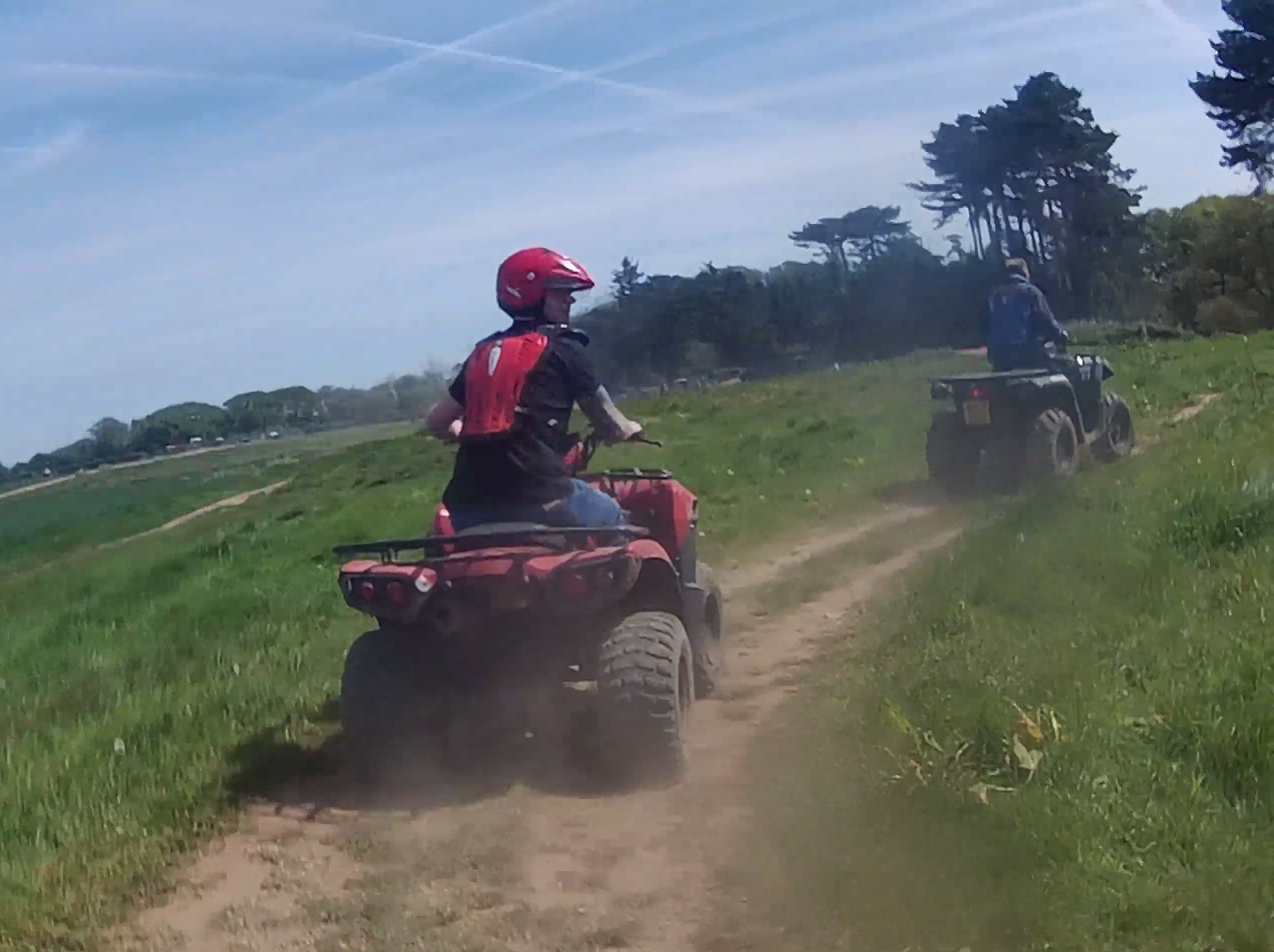 Two people riding ATVs on a dirt path through grassy fields, with one person in a red helmet in the foreground and trees in the background under a blue sky.