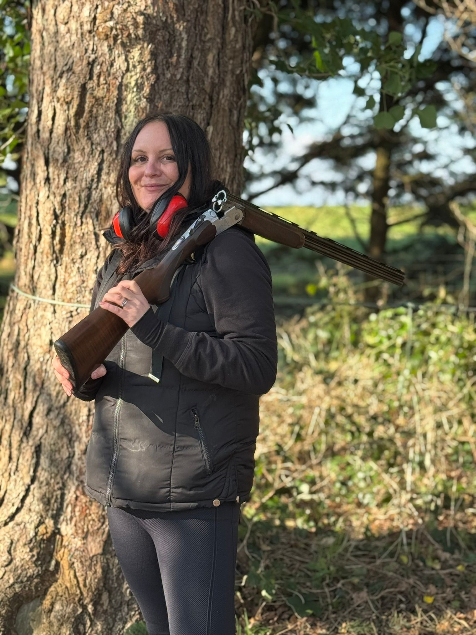 A woman standing outdoors by a tree, holding a shotgun and wearing black clothing with red earmuffs around her neck. The background shows greenery and sunlight filtering through trees.