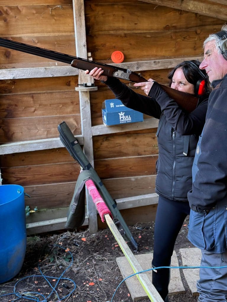 A woman wearing ear protection aims a shotgun at a clay target, with a man beside her offering guidance. They are standing in a wooden outdoor shooting range.
