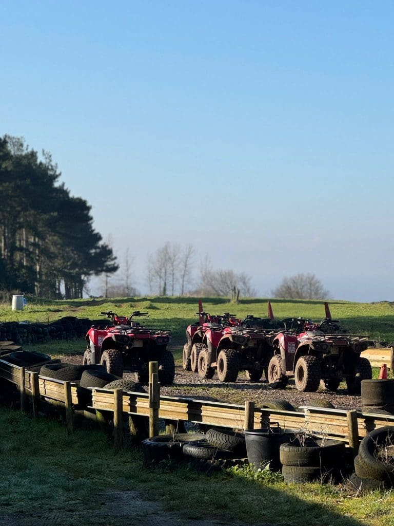 Four red ATVs are parked in a row on a dirt track lined with black tires. Green grass and a row of trees are visible in the background under a clear blue sky.
