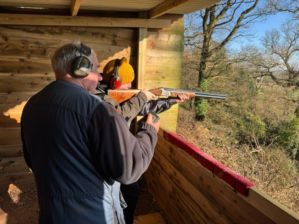 Two people stand in a wooden shooting range. The person in front, wearing a yellow hat and red earmuffs, aims a shotgun at targets outside while the instructor stands behind, guiding them. Sunlight shines through the trees outside.