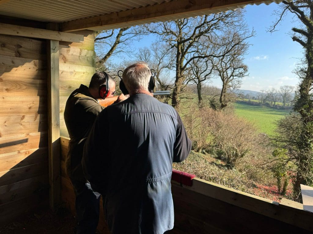 Two people wearing ear protection stand in a wooden shelter, aiming a rifle out toward a sunlit, green field with trees and blue sky in the background.