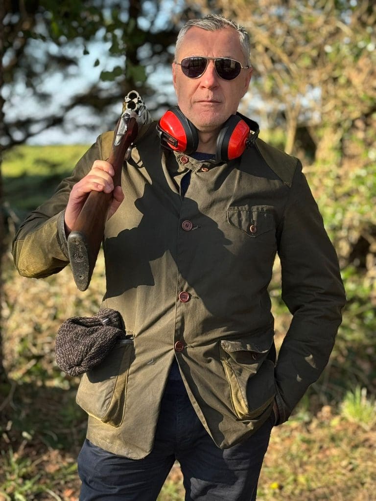 A man wearing sunglasses, an olive-green jacket, and red ear protectors around his neck stands outdoors holding a shotgun over his shoulder, with trees and sunlight in the background.