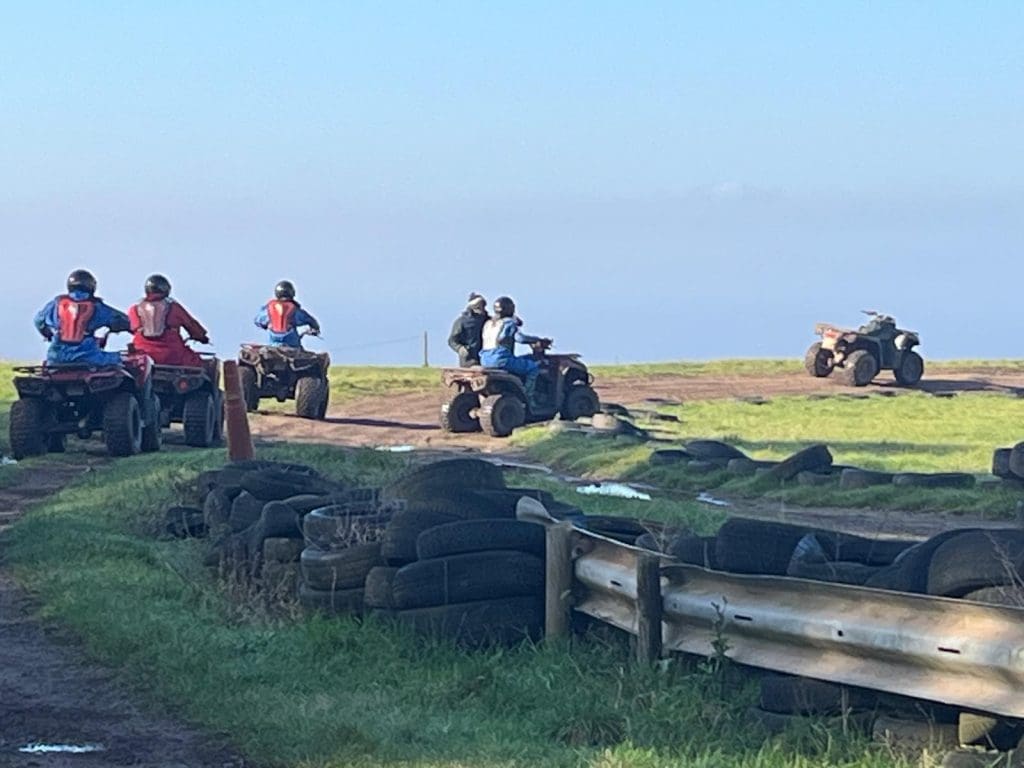 Four people ride quad bikes on a dirt track surrounded by grass, old tires, and wooden barriers under a clear sky. Another quad bike is ahead in the distance.
