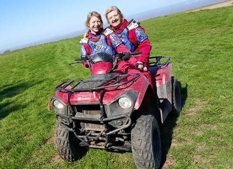 Two smiling women in red gear sit together on a red quad bike on a grassy field under a clear blue sky, enjoying an outdoor adventure.