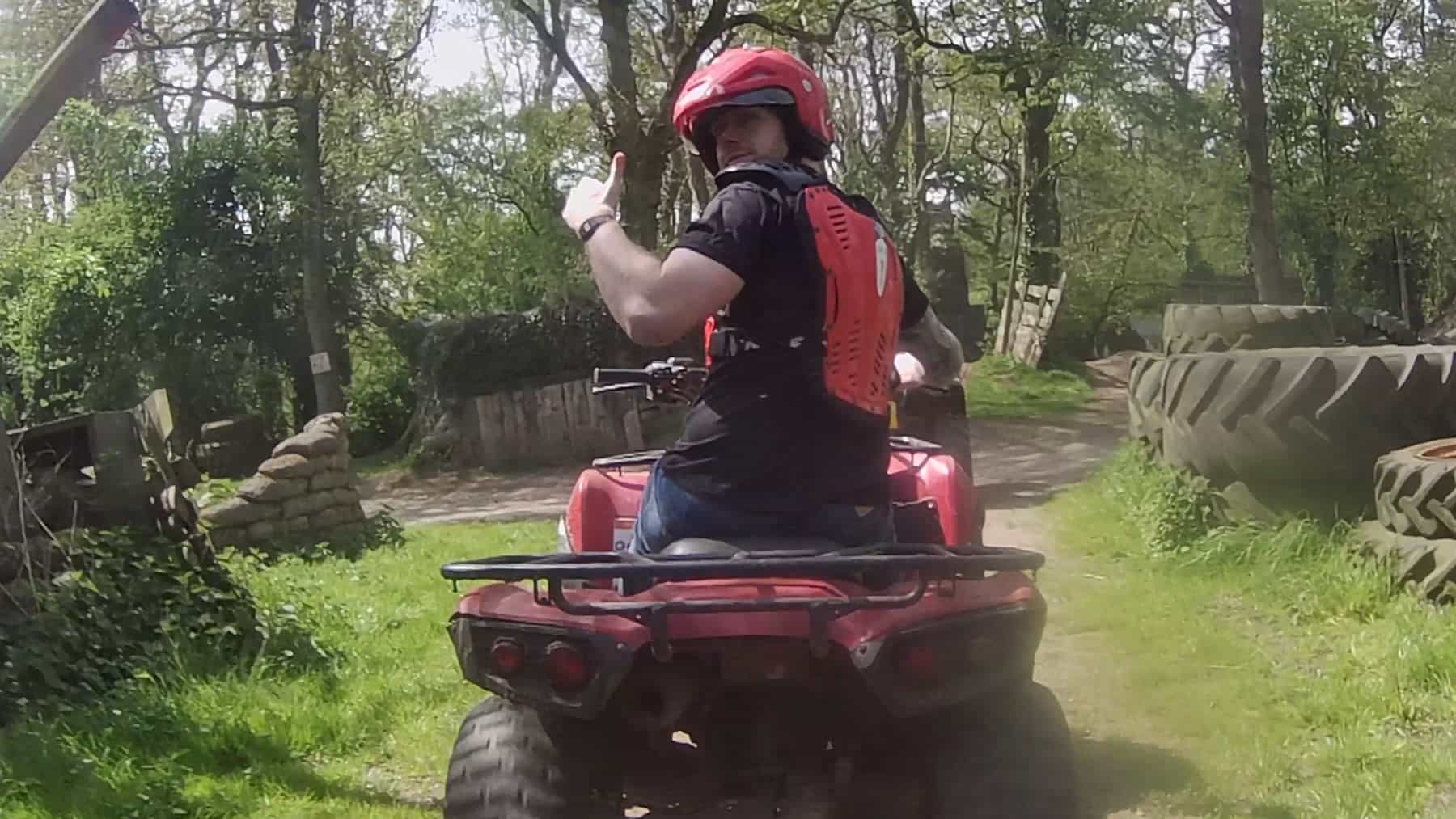 A person wearing a red helmet and protective vest rides a red ATV on a dirt path through a wooded area, looking back and giving a thumbs-up gesture. Large tires are stacked on the right side of the path.