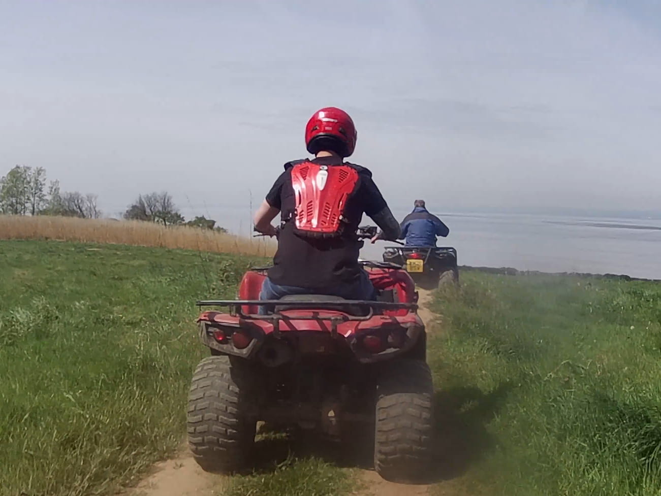 Two people ride quad bikes on a dirt path through grassy fields; the closer rider wears a red helmet and protective gear, while the other is farther ahead under a clear sky.